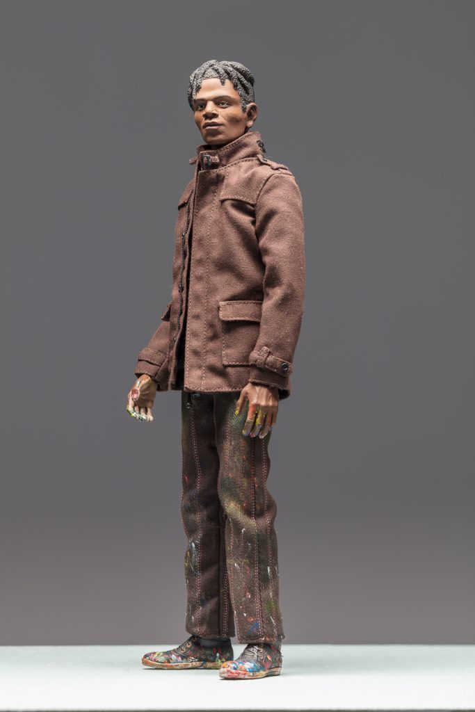 8-inch figurine of artist Jean-Michel Basquiat. He has a contemplative expression and is wearing a brown coat with his collar pulled up around his face.